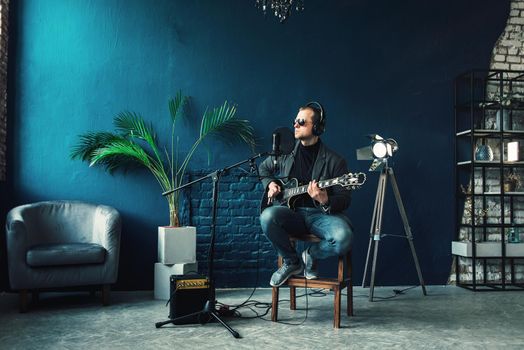 Close up of a man singer in a headphones with a guitar recording a track in a home studio. Man wearing sunglasses, jeans, black shirt and a jacket. side view