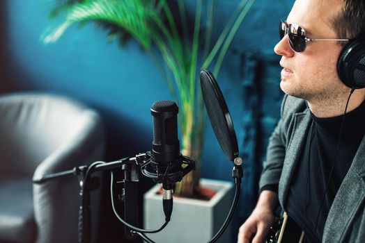 Close up of a man singer in a headphones with a guitar recording a track in a home studio. Man wearing sunglasses, jeans, black shirt and a jacket. side view