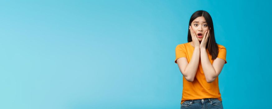 Shocked concerned young asian girl look empathy camera, gasping open mouth worried, frowning upset hear frustrating news, pitty feel sorry for friend, touch cheeks speechless, stand blue background.