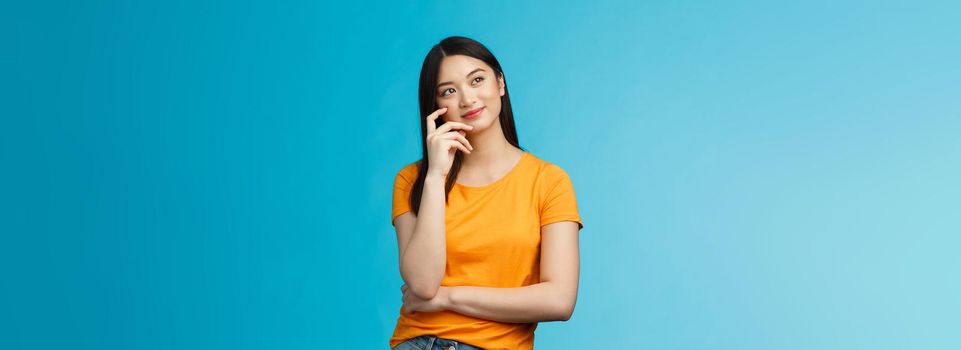 Joyful confident asian girl with dark haircut tilt head, dreaming, imaging interesting plan, smiling cunning, have idea, thinking and making choice, look up thoughtful, stand blue background.