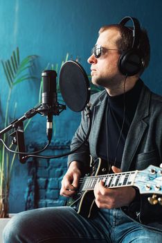 Close up of a man singer in a headphones with a guitar recording a track in a home studio. Man wearing sunglasses, jeans, black shirt and a jacket. side view