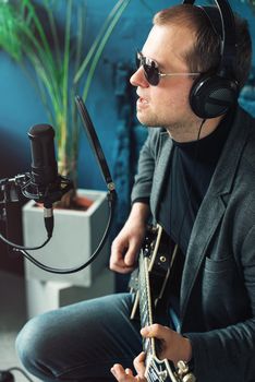 Close up of a man singer in a headphones with a guitar recording a track in a home studio. Man wearing sunglasses, jeans, black shirt and a jacket. side view