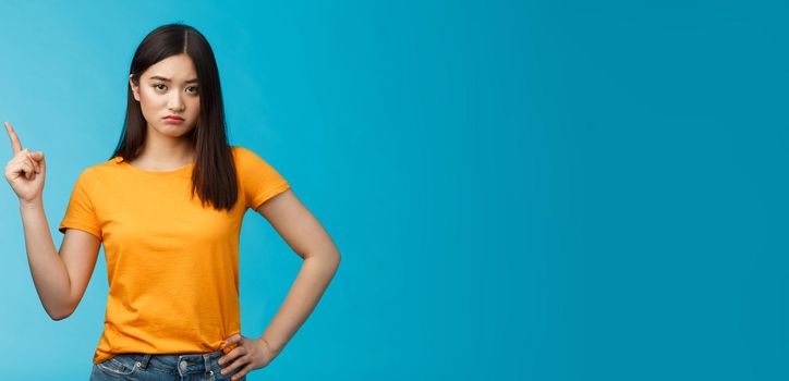 Moody upset asian girlfriend, sulking look offended and displeased pointing upper left corner, pouting disappointed, standing distressed blue background wear yellow t-shirt. Copy space