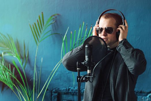 Close up of a man singer in a headphones recording a song in a home studio. Man wearing sunglasses, black shirt and a jacket. side view