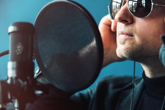 Close up of a man singer in a headphones recording a song in a home studio. Man wearing sunglasses, black shirt and a jacket. side view