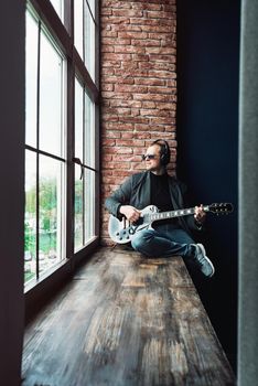 Man singer sitting on a window sill in a headphones with a guitar recording a track in a home studio. Man wearing sunglasses, jeans, black shirt and a jacket.