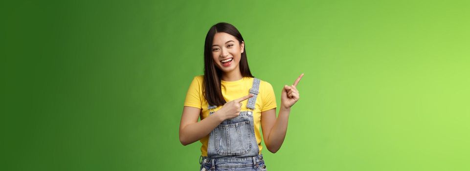 Amused friendly easygoing asian woman having fun discuss funny comedy performance, pointing left smiling laughing carefree, give positive feedback new cafe, stand green background.