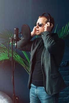 Close up of a man singer in a headphones recording a song in a home studio. Man wearing sunglasses, jeans, black shirt and a jacket. side view