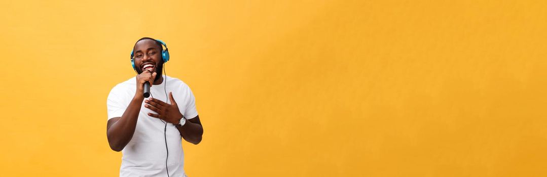 Portrait of cheerful positive chic. handsome african man holding microphone and having headphones on head listening music singing song enjoying weekend vacation isolated on yellow background.