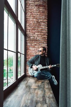 Man singer sitting on a window sill in a headphones with a guitar recording a track in a home studio. Man wearing sunglasses, jeans, black shirt and a jacket.