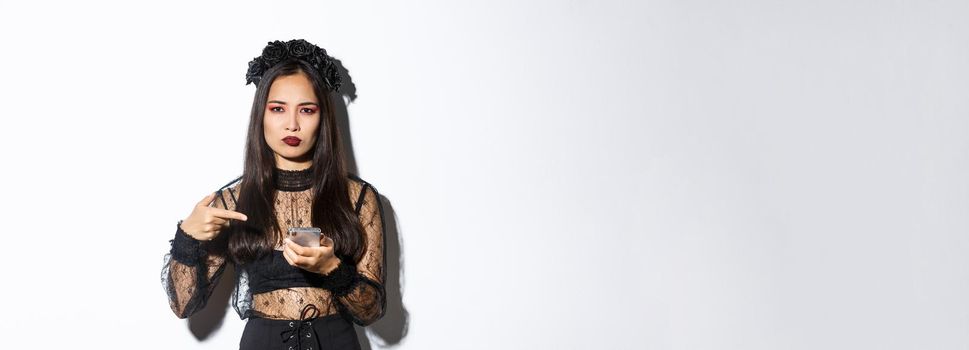 Disappointed asian woman in halloween costume, looking displeased as pointing finger at mobile phone and frowning, standing over white background.