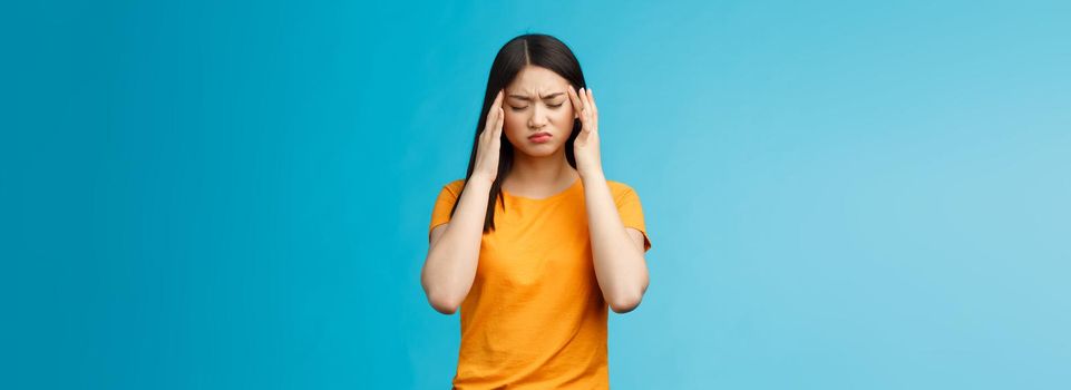 Distressed tired asian woman dark haircut cringe touch temples, suffer huge migraine need painkillers, painful headache, feel dizzy, stand blue background intense drained. Copy space