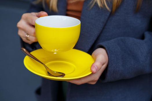 Young beautiful blonde woman in a gray coat drinks hot tea from a yellow cup on a cold autumn day. Delicious warm drink on the terrace in the cafe