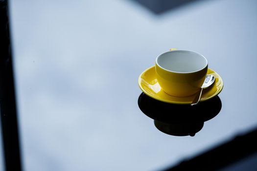 Yellow cup for coffee or tea with a saucer on a glass table.
