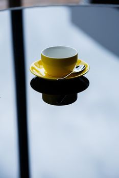 Yellow cup for coffee or tea with a saucer on a glass table.