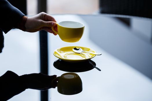 Yellow cup for coffee or tea with a saucer in the hands of a girl on the background of a glass table.
