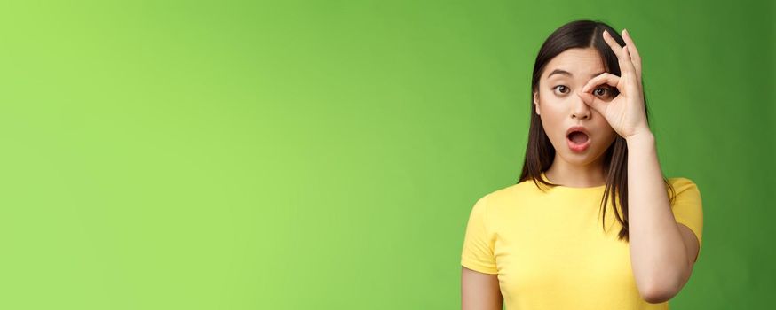 Amazed speechless cute asian young girl drop jaw astonished, look through ok circle sign, drop jaw speechless, impressed look camera, check out incredible promo, stand green background.