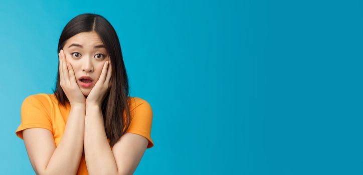 Close-up concerned shocked young timid asian girl gasping, dropping jaw feel sorry, pitty for friend open mouth stunned stare camera, grab face shook, speechless, frustrated, stand blue background.