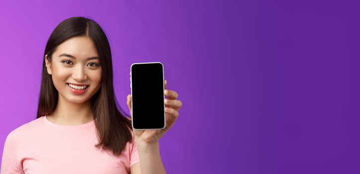 Close-up attractive pleasant female shop assistant present new phone app, hold smartphone show telephone screen, introduce application on display, stand purple background delighted.