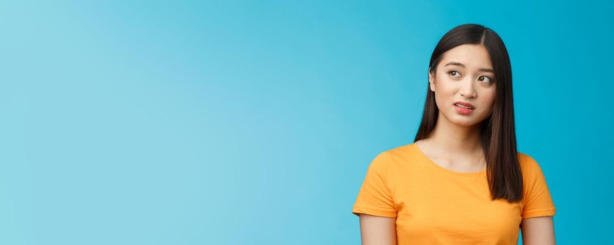 Doubtful unsure cute asian girl dark short haircut look left questioned, see strange thing, open mouth frowning upset uncertain, have hesitations, peek suspicious aside, stand blue background.