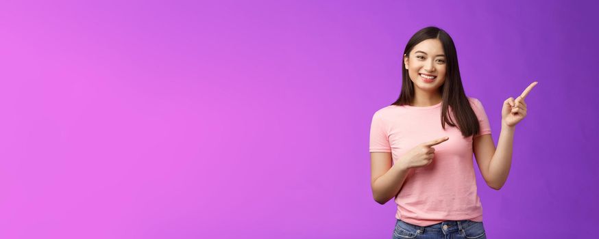 Cheerful friendly tender asian smiling girl pointing right, showing promo, introduce new product, grinning joyfully present cool adverstiment, stand pink t-shirt purple background amused outgoing.