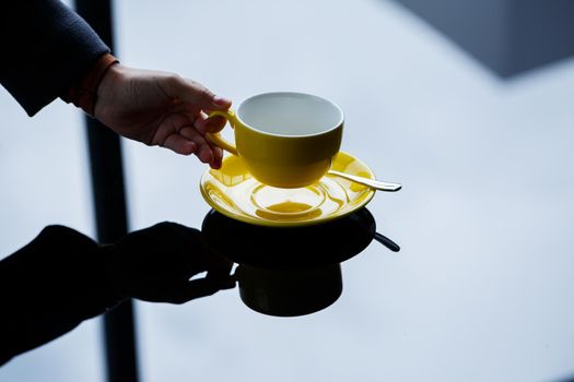 Yellow cup for coffee or tea with a saucer in the hands of a girl on the background of a glass table.