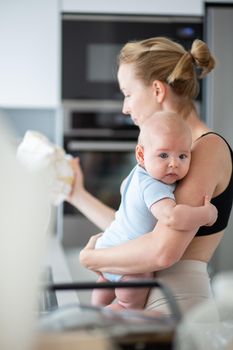 Woman wiping kitchen sink with a cloth after finishing washing the dishes while holding four months old baby boy in her hands.