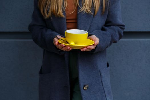 Young beautiful blonde woman in a gray coat drinks hot tea from a yellow cup on a cold autumn day. Delicious warm drink on the terrace in the cafe