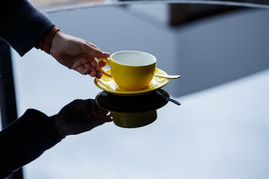 Yellow cup for coffee or tea with a saucer in the hands of a girl on the background of a glass table.