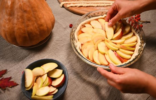 Close-up of the hands of a housewife, spreading apple slices in a circle on top of pumpkin puree in rolled out dough in the baking dish, while preparing a tasty pumpkin pie for Thanksgiving Day