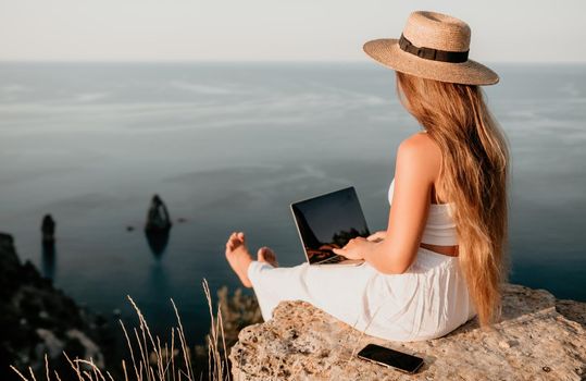 Successful business woman in yellow hat working on laptop by the sea. Pretty lady typing on computer at summer day outdoors. Freelance, travel and holidays concept.