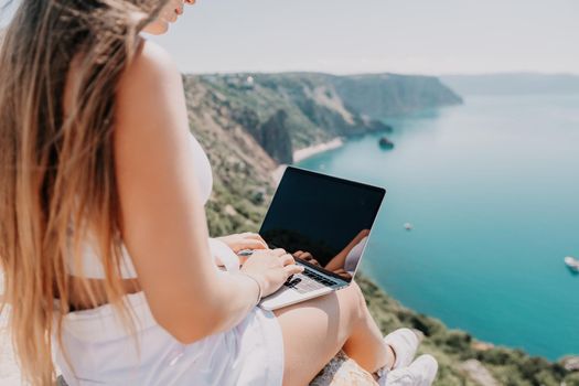 Successful business woman in yellow hat working on laptop by the sea. Pretty lady typing on computer at summer day outdoors. Freelance, travel and holidays concept.