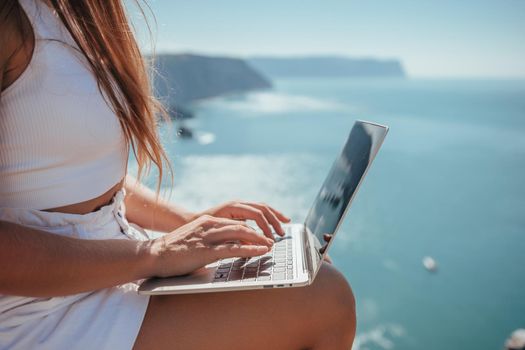 Successful business woman in yellow hat working on laptop by the sea. Pretty lady typing on computer at summer day outdoors. Freelance, travel and holidays concept.