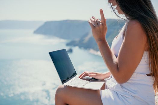 Successful business woman in yellow hat working on laptop by the sea. Pretty lady typing on computer at summer day outdoors. Freelance, travel and holidays concept.