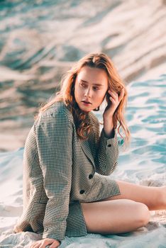 Debonair blonde girl sitting at sandy beach in autumn sunset. Outdoor portrait of pretty curly fashionable girl
