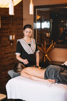 Specialist making biolocation with crystal pendulum. Young charming girl on a panchakarma procedure laying on a massage table. beautiful woman spending time at modern spa cabinet relaxing. Soft yellow light.