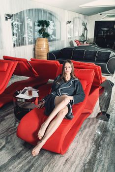 Portrait of young beautiful woman with long sexy legs relaxing in a fashionable red chair in a gray bathrobe, waiting for treatment. Luxery spa center