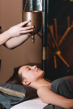 Specialist making shirodhara massage form of ayurveda therapy. Young charming girl on a panchakarma procedure laying on a massage table. beautiful woman spending time at modern spa cabinet relaxing. Soft yellow light.