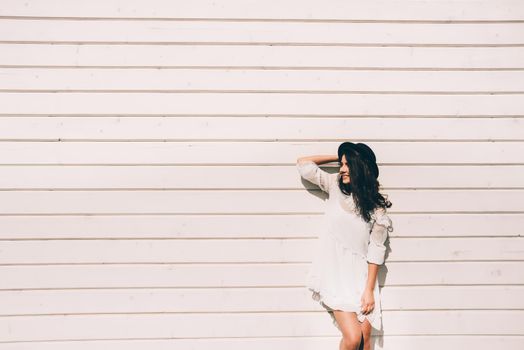 Sunny lifestyle fashion portrait of young stylish hipster woman walking on the street, wearing trendy white dress, black hat and boots. White wooden backgrond.