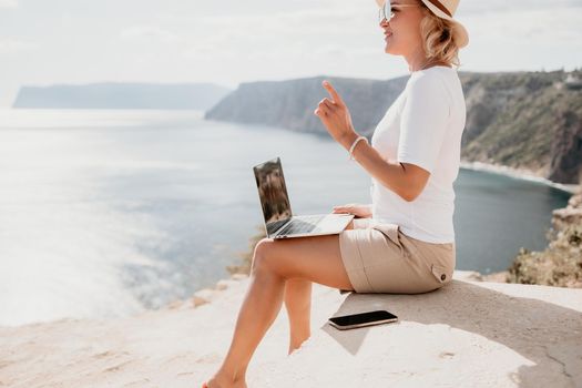Successful business woman in yellow hat working on laptop by the sea. Pretty lady typing on computer at summer day outdoors. Freelance, travel and holidays concept.