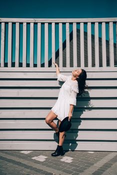 Sunny lifestyle fashion portrait of young stylish hipster woman walking on the street, wearing trendy white dress, black hat and boots. Gray wooden backgrond.