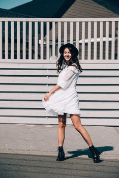 Sunny lifestyle fashion portrait of young stylish hipster woman walking on the street, wearing trendy white dress, black hat and boots. Gray wooden backgrond.