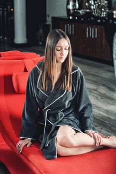 Portrait of young beautiful woman with long sexy legs relaxing in a fashionable red chair in a gray bathrobe, waiting for treatment. Luxery spa center