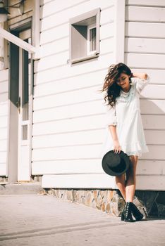 Sunny lifestyle fashion portrait of young stylish hipster woman walking on the street, wearing trendy white dress, black hat and boots. White wooden house on a backgrond.