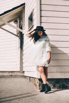 Sunny lifestyle fashion portrait of young stylish hipster woman walking on the street, wearing trendy white dress, black hat and boots. White wooden house on a backgrond.
