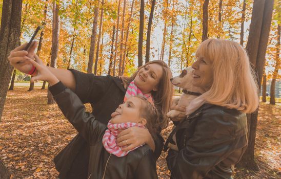 Three generations of women and dog feel fun look at camera posing for self-portrait picture together, funny excited child, mom and grandmother have fun enjoy weekend take selfie on gadget on autumn park