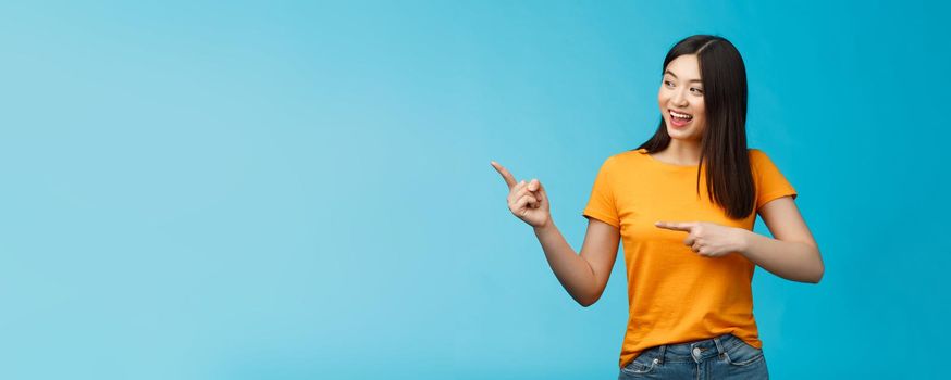 Friendly enthusiastic asian girl curiously looking sideways, pointing left interesting object, grinning joyfully discuss new opened store, stand blue background carefree lively talking.