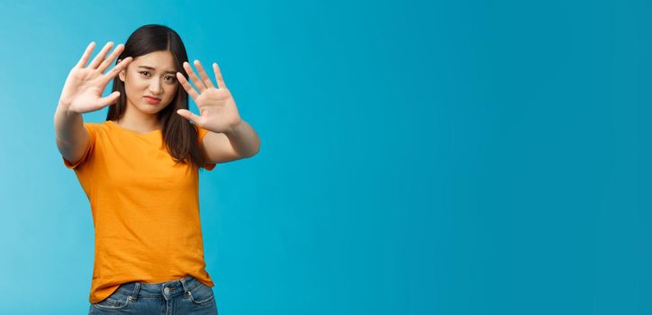 Asian moody displeased silly timid girl asking turn off light not photograph her, raise hands block, cover face step back and cringe disappointed, standing blue background upset reluctant. Copy space