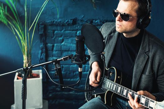 Close up of a man singer in a headphones with a guitar recording a track in a home studio. Man wearing sunglasses, jeans, black shirt and a jacket. side view