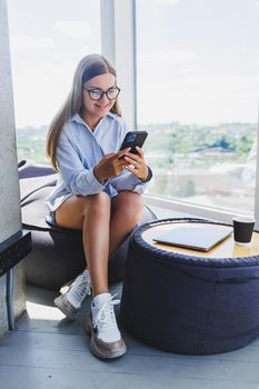 Happy freelancer girl in classic glasses looking at phone and smiling while sitting in modern coworking space, carefree millennial woman in glasses enjoying leisure time for communication